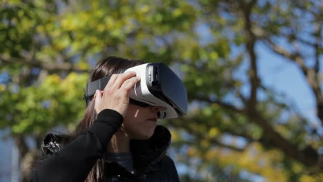 Young-dark-haired-woman-in-warm-black-waistcoat-wearing-virtual-reality-glasses-in-early-autumn-park